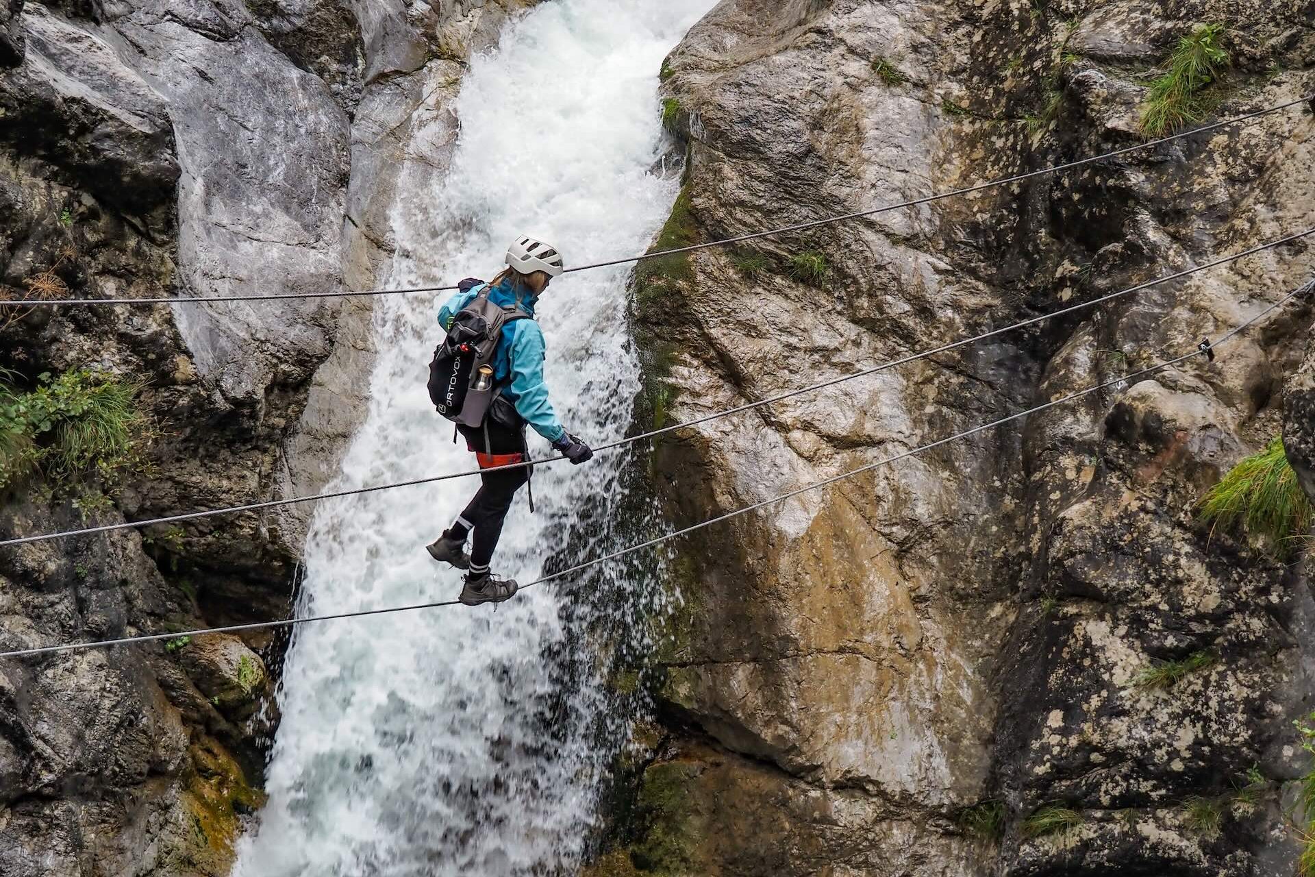 Frau mit Helm und KlettersteigausrÃ¼stung balanciert auf einer SeilbrÃ¼cke, die Ã¼ber einen Wasserfall verlÃ¤uft.